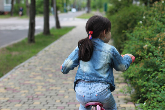 Little girl in a denim suit rides a pink bike along a brick multi-colored path in a spring city