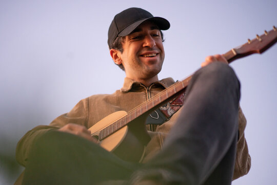 Hombre afeitado con gorra negra y cazadora de pana tocando la guitarra al aire libre con el cielo despejado de fondo, sonriendo