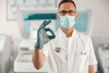 Young doctor with rubber gloves and face mask standing in hospital and showing okay. Corona virus is going down.