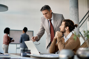 Mature business mentor reviews work of his younger colleague on laptop in the office.