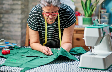 Vertical photo of woman with white hair, wearing glasses and sewing tape around her neck, working at the sewing table at home.