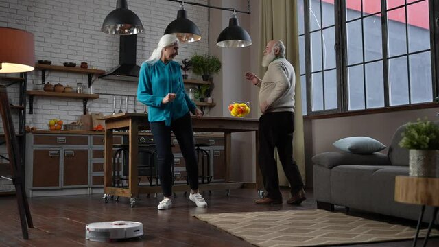 Cheerful aged gray-haired couple enjoying joint dance in free time while robotic vacuum cleaner working. Positive modern elderly family having fun in dance while automatic cleaner washing floor
