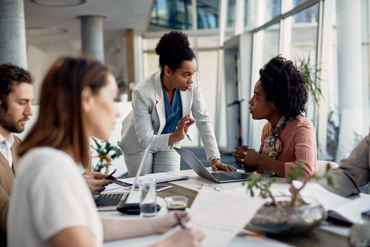 Female business mentor talks to assistant manager who works on laptop during meeting in the office.