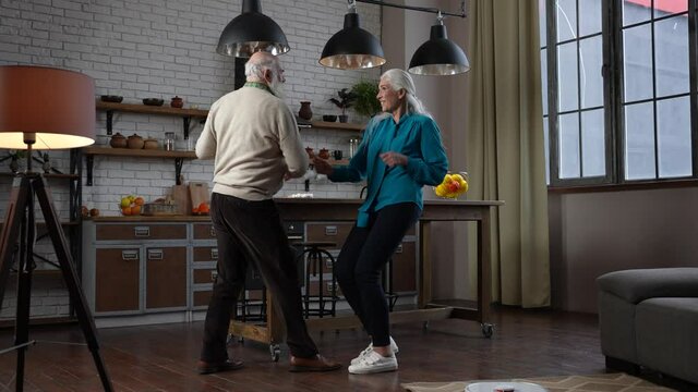Cheerful elderly white-haired people having fun in free time while robot vacuum cleaner working. Active modern aged family spending domestic leisure with pleasure while automatic hoover washing floor