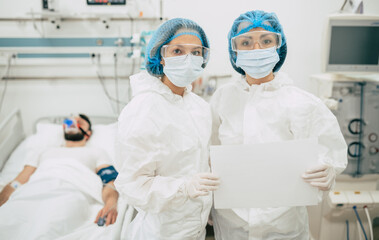 Two confident doctors in protective safety suits standing on background of a patient with...