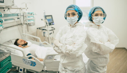 Two confident doctors in protective safety suits standing on background of a patient with...