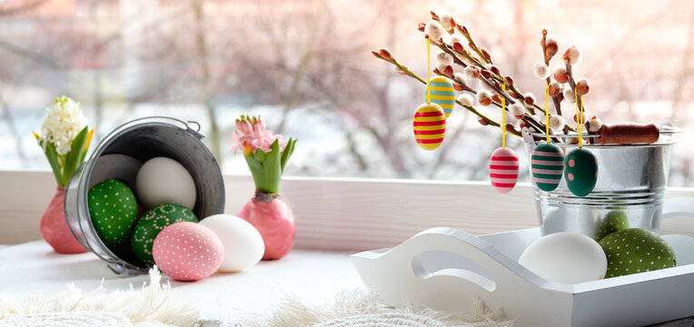 Easter decorations on windowsill in Spring. Wooden painted eggs on pussy willow twigs in glass bottle, Easter eggs in rattan basket, rustic bucket, deco tray. Window with sunset light, view over town.