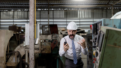 Fototapeta na wymiar A beard mechanical engineer, supervisor or factory manager is communicating with his staff by using radio or handhold walkie talkie in a warehouse. Smart manager with helmet is getting mad in mission