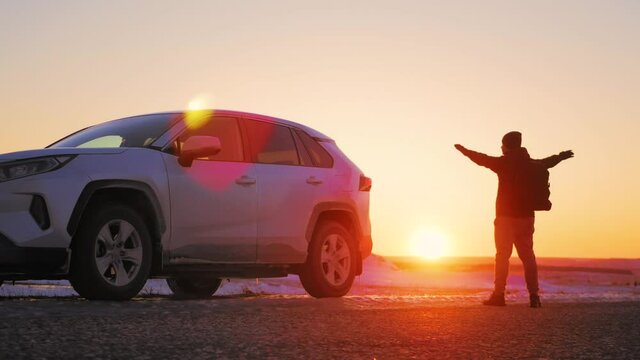 Back view of a silhouette man with backpack arms wide open to the sides at sunset. Young male traveler is enjoying beautiful landscape during road trip on suv. Road trip, travel and people concept.