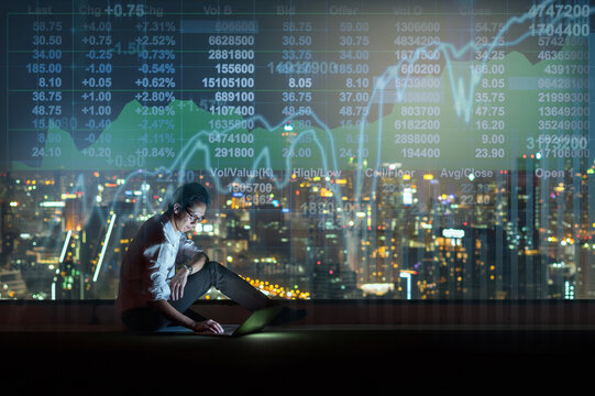 Asian businessman sitting and using the smart mobile phone showing the Stock market chart over the cityscape background at night time, Business  technology and trading concept