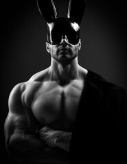 Portrait of naked shirtless muscular man with perfect built body wearing black glossy rabbit mask standing, showing biceps and chest, holding formal jacket on his shoulder over dark background