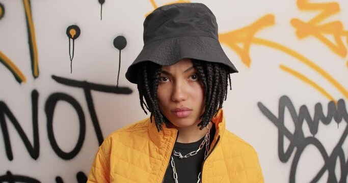 Urban subculture concept. Stylish cheeky woman wears trendy hat yellow jacket poses against graffiti wall has cheeky expression. Female rocker spends free time in circle of friends. Bad street girl