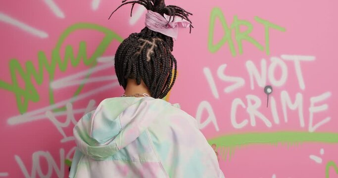 Youth subculture art and creativity concept. Hipster hip hop girl with combed braids draws colorful graffiti with aerosol spray on steet wall dressed in white hoodie demonstrated her talents