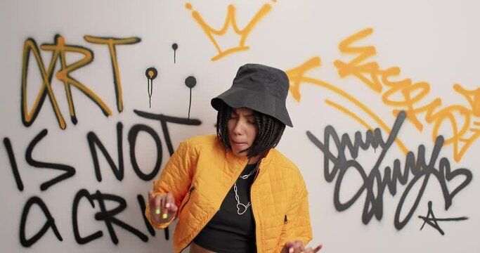 Youth and outdoor lifestyle concept. Happy gangster girl in fashionable clothes moves actively raises arms dances against graffiti wall in urban environment has fun. Trendy female steet artist