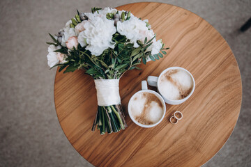 Wedding morning. A wedding bouquet, two cups of coffee and wedding rings lie on the table. aesthetics and details of the wedding - 421981899