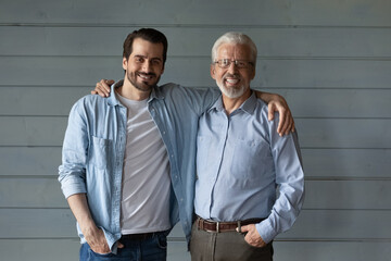 Portrait of smiling old Caucasian 60s father and adult millennial son stand isolated on grey wall...