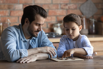 Loving young Caucasian father and little 7s son relax at home play checkers or draughts. Caring 30s...