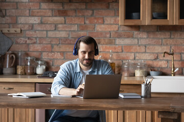 Focused young Caucasian man in headphones work online on computer at home kitchen. Millennial male...