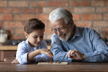 Smiling senior 60s grandfather sit at table have fun play checkers draughts with small 7s grandson. Happy older granddad teach little smart grandchild, engaged in logical set or game at home together.