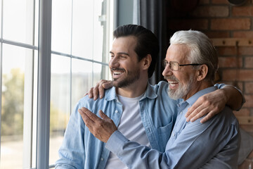 Fototapeta na wymiar Smiling senior grey-haired Caucasian dad and adult son look in window distance thinking planning together. Happy mature 60s father and grownup man child enjoy family weekend, dream or visualize.