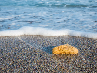 A rock on the beach in the sand with the water of the foamy sea in the background.