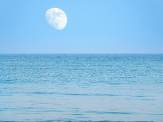 The moon in the blue sky hanging above the water of the sea or ocean.