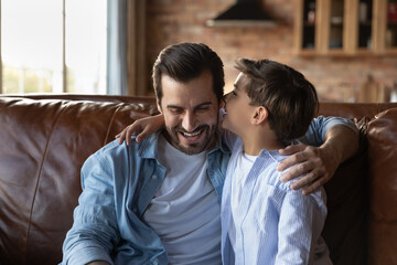 Cute little 7s Caucasian boy kid whisper tell secret to excited young father, relaxing together at home. Small happy son child have fun with loving caring single dad in living room. Family concept.