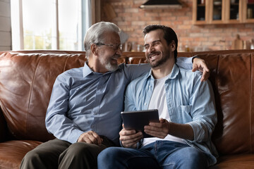 Overjoyed Caucasian adult man child relax on sofa at home with mature modern dad use tablet together. Smiling young adult grownup son hold pad rest on couch in living room with elderly father.