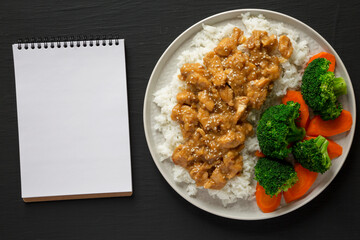 Homemade Orange Chicken with White Rice, Broccoli and Carrot, blank notepad on a black surface, top view. Flat lay, overhead, from above.