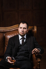 a bearded man sits in luxury leather armchair and relax with cigar and a glass of whiskey