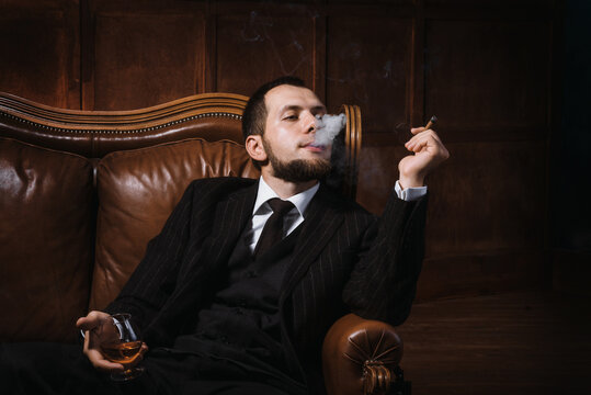 a bearded man sits in luxury leather armchair and relax with cigar and a glass of whiskey