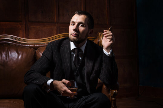a bearded man sits in luxury leather armchair and relax with cigar and a glass of whiskey