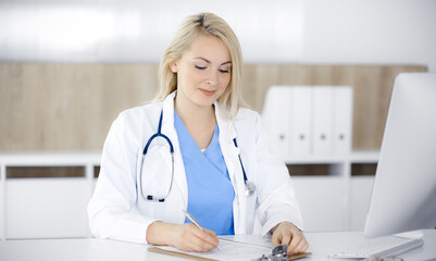 Woman-doctor at work while sitting at the desk in hospital or clinic. Blonde cheerful physician filling up medication history record form