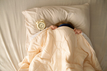 In the early morning, a preschool girl lies in bed under the covers next to the alarm clock. The...