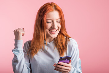 Fototapeta premium Redhead girl looks at the phone and shows the gesture of the winner in the studio on a pink background