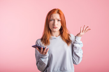 Confused redhead girl with phone in studio on pink background