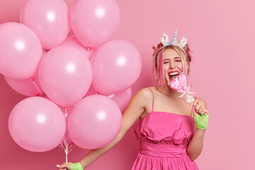 Cheerful blonde woman has party mood bites heart shaped lollipop keeps mouth wide opened wears stylish dress holds bunch of inflated balloons isolated over pink background. Birthday celebration