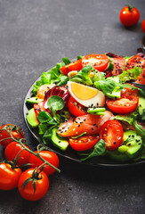 Keto diet salad with salted salmon, avocado, egg, sesame seeds, olive oil, tomatoes and mixed herbs. Black background, top view, copy space