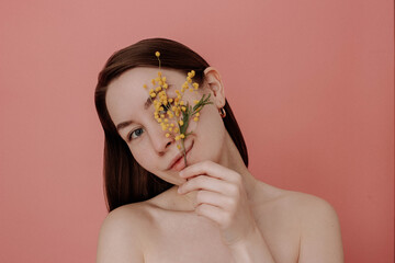 Smiling young woman with naked shoulders and no makeup holding branch of yellow mimosa flower on pink background studio shot. Smiling girl with brown hair. Spring, summer concept. Natural beauty