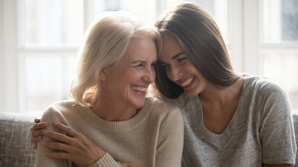 Overjoyed millennial Caucasian woman embrace happy older 60s mom enjoy family weekend reunion together. Smiling mature mother and adult grownup daughter hug laugh sharing secrets at home. - obrazy, fototapety, plakaty