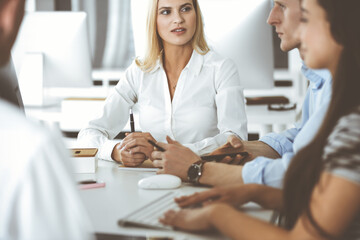 Fototapeta premium Group of business people discussing questions at meeting. Headshot of blonde businesswoman while smiling to her colleague at office negotiation. Teamwork and cooperation in corporate occupation