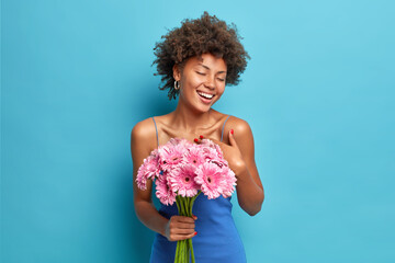 Happy dark skinned pleased woman enjoys romantic date gets bouquet of gerbera flowers from boyfriend closes eyes from pleasure isolated over blue background feels very thankful. Feminity concept - Powered by Adobe