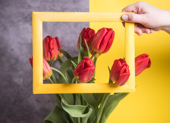 A woman's hand holds a yellow frame over tulips arranged on a Ultimate Gray-yellow background. The trend background of 2021.