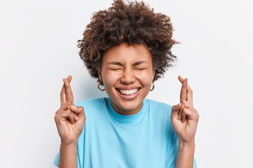 Close up shot of curly haired happy diligent female student makes wish before exam awaits dreams come true crosses fingers smiles positively wears casual blue clothes isolated over white background