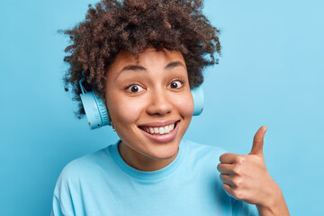 Portrait of curly haired Afro American listens music enjoys favorite track via headphones keeps thumb up shows excellent sign smiles happily isolated over blue background. Body language concept