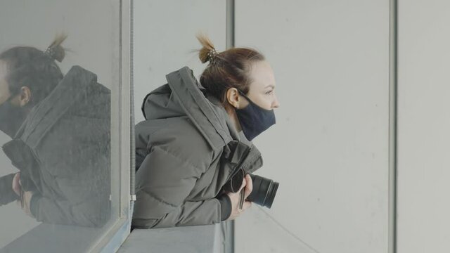 Female photographer looks out of window with camera. Action. Masked paparazzi woman with camera looks out of window. Side view of woman looking out of window