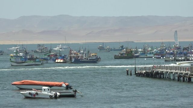 Remote port with sandy beaches in Paracas, Peru. White motor-boat passes with high speed between a pier and a row of anchored fishing boats.