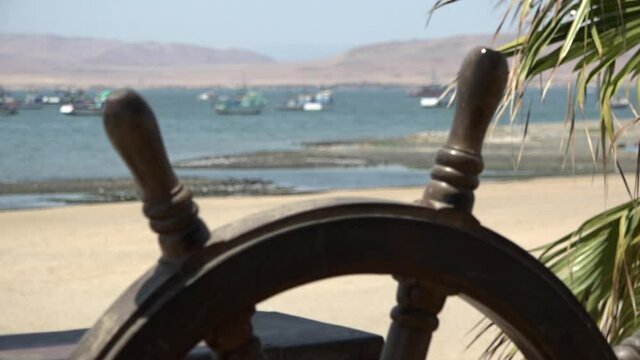 Steering wheel of an old-fashioned ship with palms, sandy beach and a harbor at the background. Paracas, Peru.