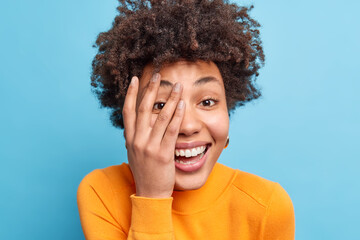 Close up shot of positive dark skinned pretty woman smiles broadly covers face with hand expresses...
