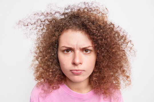 Close up portrait of sullen curly haired woman smirks face has offended troubled suspicious expression satres at camera with dissatisfaction isolated over white background. Emotions concept.